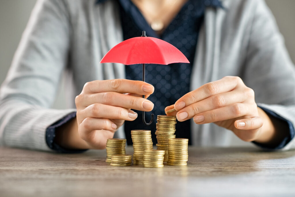 Young woman hands holding red umbrella over stacked coin on table. Female hand holding a small umbrella to protect heaps of coins while saving them. Financial security and savings protection concept.